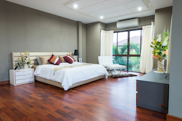 a large room with a hardwood floor, a tall window overlooking tress, a two-person bed with a single bedside able, and a padded chair next to the window